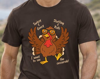 Może przedstawiać: Ciemnobrązowy t-shirt z krótkim rękawem z grafiką indyka w okularach z soczewkami w kształcie ciasta. Napis głosi: Turkey & Gravy, Stuffing & Rolls, I want to see the casserole! Indyk ma rozpostarte skrzydła i czerwoną narośl na szyi.