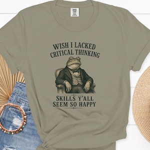 May include: A sage green short-sleeve t-shirt featuring a graphic of a frog wearing a suit and bowtie while sitting in a chair. The text above the frog reads WISH I LACKED CRITICAL THINKING and the text below reads SKILLS Y'ALL SEEM SO HAPPY.
