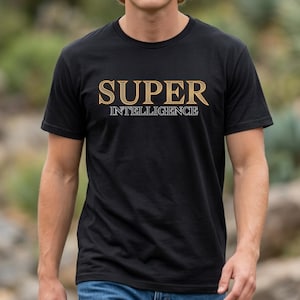 Puede incluir: Una camiseta negra de manga corta con el texto SUPER en letras grandes de tono dorado con un contorno oscuro, situado sobre la palabra INTELLIGENCE en letras más pequeñas con contorno blanco. La camiseta tiene cuello redondo.