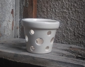 May include: White ceramic planter with a round shape and multiple circular holes. The planter is sitting on a weathered wooden surface.