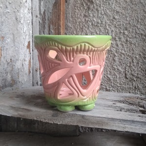 May include: A ceramic planter with a pink body and a green rim. The planter features cut-out designs and sits on three small feet. The background shows weathered wood and a concrete wall.
