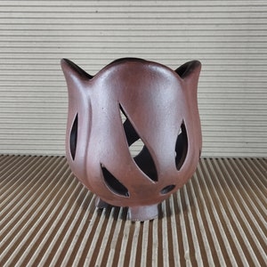 May include: A dark brown ceramic vessel with a smooth, matte finish and a tulip-like shape. The body features several leaf-shaped cutouts throughout its surface, and the base is supported by three small, integrated feet for stability.