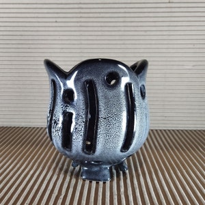 Może przedstawiać: Ceramiczny świecznik o zaokrąglonym, pękatym kształcie i błyszczącym wykończeniu. Powierzchnia posiada gradient czerni i bieli z nakrapianą teksturą. Posiada pionowe wycięcia oraz małe okrągłe otwory i wspiera się na trzech krótkich nóżkach.