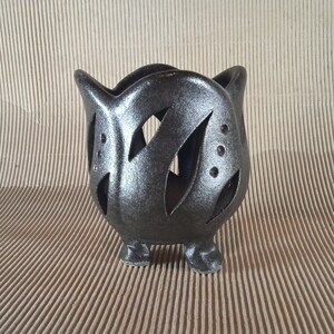 May include: A dark grey ceramic candle holder with a distinctive design. The holder has a rounded shape with cut-out leaf-like patterns and small circular holes. It stands on three small feet, creating a decorative and functional piece.