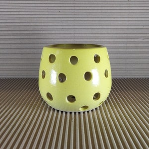 May include: A light yellow ceramic candle holder with multiple circular holes. The holes are evenly spaced around the sides of the holder. The interior is a darker shade of yellow. The candle holder is on a corrugated surface.