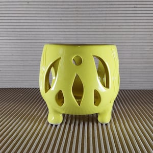May include: A bright yellow ceramic candle holder featuring a rounded body with intricate cut-out patterns and three small feet. The surface has a smooth, glossy finish with subtle vertical embossed lines along the sides of the vessel.