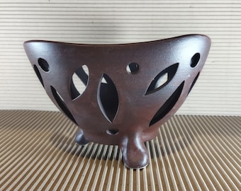 May include: A brown ceramic bowl with a distinctive design. The bowl has a curved shape and is supported by three small feet. It features decorative cut-out leaf and circular patterns around the sides. The bowl is likely used for decorative purposes.