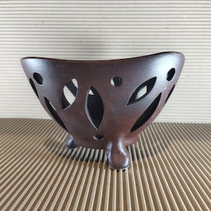 May include: A brown ceramic bowl with a distinctive design. The bowl has a curved shape and is supported by three small feet. It features decorative cut-out leaf and circular patterns around the sides. The bowl is likely used for decorative purposes.