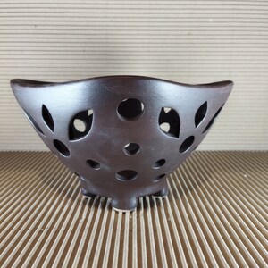 Puede incluir: Un cuenco de cerámica marrón oscuro con un diseño único. El cuenco presenta una serie de recortes circulares y en forma de hoja en los laterales. Tiene un borde ancho, ligeramente ondulado y descansa sobre tres pequeños pies. El cuenco probablemente está destinado a fines decorativos o de exhibición.