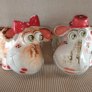 May include: Two ceramic figurines of cartoon-style animals with large eyes. The figurine on the left is brown and white with a red bow. The figurine on the right is white with a red hat.