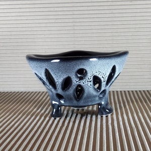 May include: A ceramic planter with a wide, flared rim and three short legs. The exterior features a black and white speckled glaze with a series of oval-shaped cutouts distributed around the body, revealing the dark interior of the vessel.