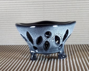 May include: A ceramic planter with a wide, flared rim and three short legs. The exterior features a black and white speckled glaze with a series of oval-shaped cutouts distributed around the body, revealing the dark interior of the vessel.
