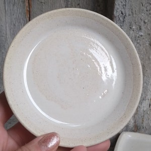 May include: A small, white ceramic dish with a speckled glaze. The dish is round and has a slightly raised rim.