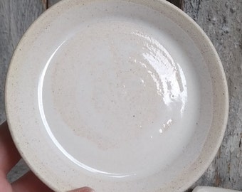 May include: A small, white ceramic dish with a speckled glaze. The dish is round and has a slightly raised rim.