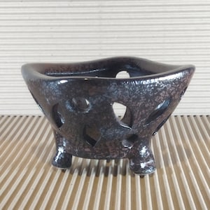 May include: A black ceramic planter with a unique, irregular shape and a three-legged base. The planter has a speckled glaze and intricate cutouts around the sides.
