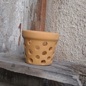 May include: A terracotta planter with a circular pattern of holes. The planter is a light brown colour and is sitting on a wooden surface.