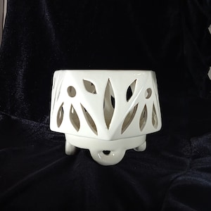 May include: White ceramic planter with a geometric design and three legs. The planter has cut-out details in the shape of leaves.