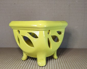 May include: A bright yellow ceramic planter with a square rim and three short, sturdy feet. The sides of the planter feature decorative cutouts in a leaf-like pattern, allowing for visibility through the body of the vessel to the interior.