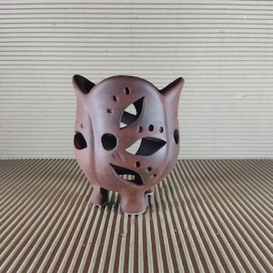 Może przedstawiać: Brązowy ceramiczny świecznik w kształcie sowy z wyciętymi wzorami oczu i piór. Świecznik ma trzy małe nóżki i dwa spiczaste uszy. Powierzchnia ma matowe wykończenie i jest umieszczona na tle z tektury falistej.