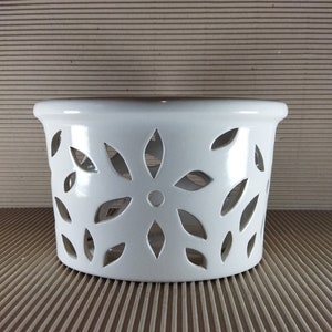 Peut inclure: Un récipient en céramique blanche avec un motif floral découpé. Le récipient est de forme cylindrique avec un bord légèrement évasé. Les découpes ont la forme de feuilles, disposées en motif floral sur les côtés.