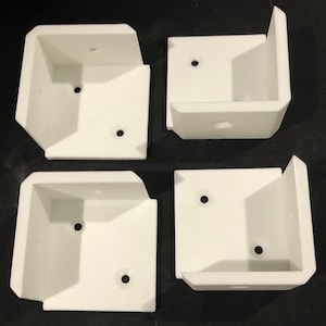 May include: Four white, right-angled brackets, each with two pre-drilled holes. The brackets are made of a solid material and have a clean, modern design. They are likely used for shelving or support.