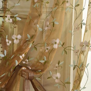 Könnte beinhalten: Durchsichtige braune Vorhänge mit gestickten weißen Blumen und grünen Blättern.