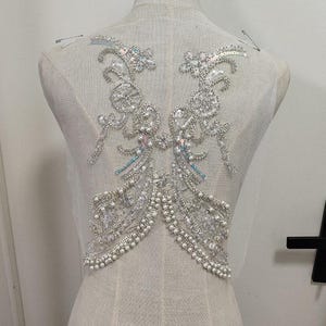May include: A silver and white beaded applique with a floral design. The applique is on a sheer fabric background.