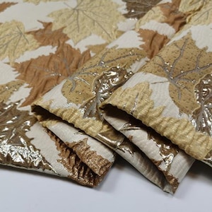 Può includere: Primo piano di un tessuto con un motivo a foglie dorate e marroni. Il tessuto è piegato in tre parti e ha una superficie testurizzata.