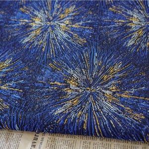May include: A dark blue fabric featuring a repeating pattern of starburst or firework motifs in shades of gold, light blue, and white. The surface has a textured, pleated appearance with radiating lines extending from the center of each design.