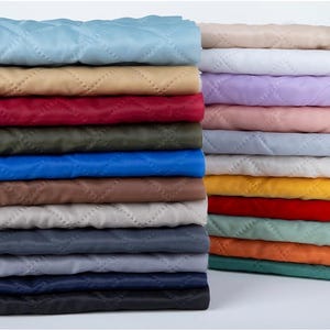 May include: A stack of folded quilted fabric in a variety of solid colours including light blue, tan, red, olive green, royal blue, brown, light grey, dark grey, black, cream, white, lavender, pink, yellow, orange, and teal with diamond stitching.