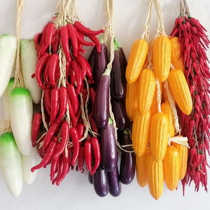 Pu&ograve; includere: Assortimento di verdure artificiali, tra cui peperoncini rossi, melanzane viola, mais giallo in pannocchia e ravanelli bianchi. Le verdure sono legate con lo spago e pendono da uno sfondo bianco.