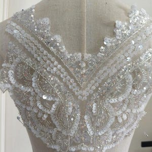 Może przedstawiać: Biała aplikacja z koralików i cekinów z delikatnym wzorem kwiatowym. Aplikacja idealnie nadaje się do dodania blasku sukien ślubnych, sukien wieczorowych lub innych strojów na specjalne okazje.