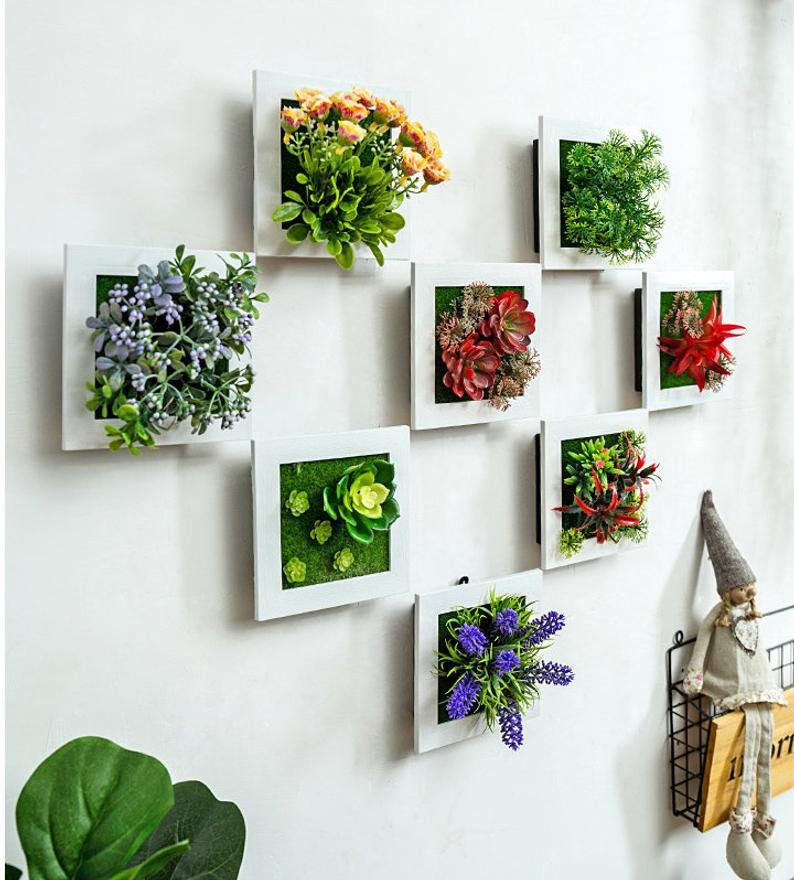 Puede incluir: Nueve marcos cuadrados blancos con diferentes plantas y flores artificiales en su interior. Los marcos est&aacute;n dispuestos en forma de cuadr&iacute;cula en una pared blanca. Las plantas incluyen suculentas, lavanda y helechos.