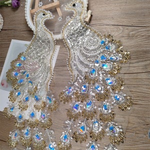 May include: Two white peacock embroidery patches with iridescent blue and gold sequins. The patches are on a white mesh background.