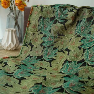 Pode incluir: Close-up de um tecido com um padrão de folhas repetido. As folhas são em tons de verde e dourado, com detalhes em vermelho, sobre um fundo preto. O tecido tem uma aparência texturizada, sugerindo um material tecido.
