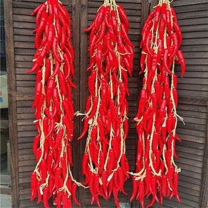Pu&ograve; includere: Tre fili di peperoncini rossi artificiali appesi a una parete di legno. I peperoncini sono legati insieme con lo spago e sono disposti in modo decorativo.