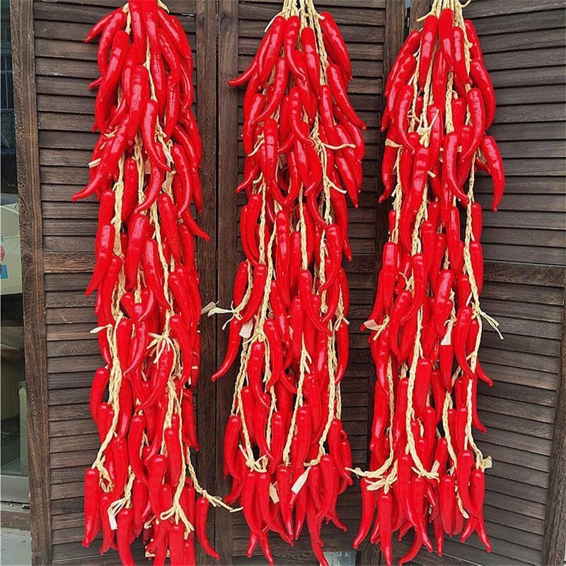 Pu&ograve; includere: Tre fili di peperoncini rossi artificiali appesi a una parete di legno. I peperoncini sono legati insieme con lo spago e sono disposti in modo decorativo.
