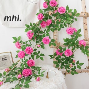 May include: Artificial pink rose vine with green leaves, hanging from a wooden ladder. The vine is displayed on a white fuzzy surface with a white bag that says "mhl." and a photo frame.