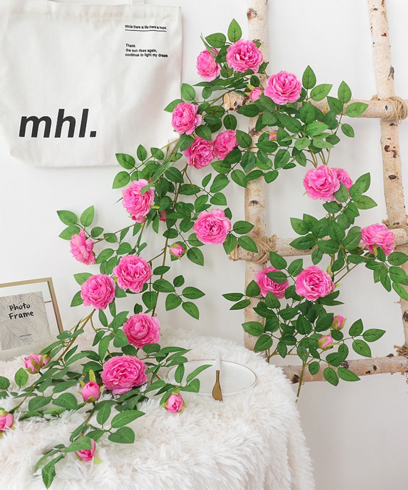 May include: Artificial pink rose vine with green leaves, hanging from a wooden ladder. The vine is displayed on a white fuzzy surface with a white bag that says "mhl." and a photo frame.