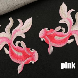 Könnte beinhalten: Zwei rosa bestickte Goldfische mit weißen Akzenten. Die Goldfische befinden sich auf einem schwarzen Hintergrund. Das Wort "rosa" steht unter den Goldfischen.