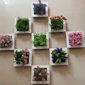 Puede incluir: Nueve marcos cuadrados blancos con diferentes plantas y flores artificiales. Los marcos est&aacute;n dispuestos en una cuadr&iacute;cula en una pared blanca. Las plantas y flores incluyen lavanda, hierba verde, flores rosas y flores moradas.