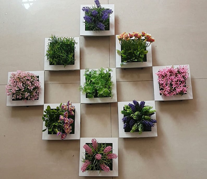 Puede incluir: Nueve marcos cuadrados blancos con diferentes plantas y flores artificiales. Los marcos est&aacute;n dispuestos en una cuadr&iacute;cula en una pared blanca. Las plantas y flores incluyen lavanda, hierba verde, flores rosas y flores moradas.