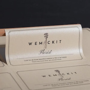 以下が含まれることがあります： WEMCKIT Floristというテキスト、花を持つ手のイラスト、そして「Look for all those beautiful reasons in life to be happy. There are more than you would think.」という引用文が繰り返された、長方形のベージュ色のステッカーシート。