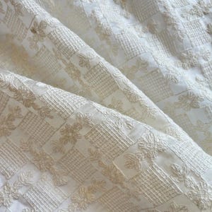 May include: A white fabric featuring a repeating pattern of embroidered beige floral motifs and square grid sections. The material is gathered into soft folds, highlighting the texture of the intricate needlework and the contrast between the light base and the tan thread.