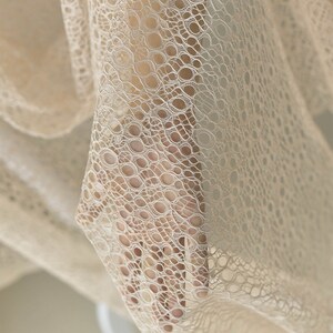May include: Close-up of a beige lace fabric with a delicate, circular pattern. The fabric is soft and flowing, with a subtle sheen.