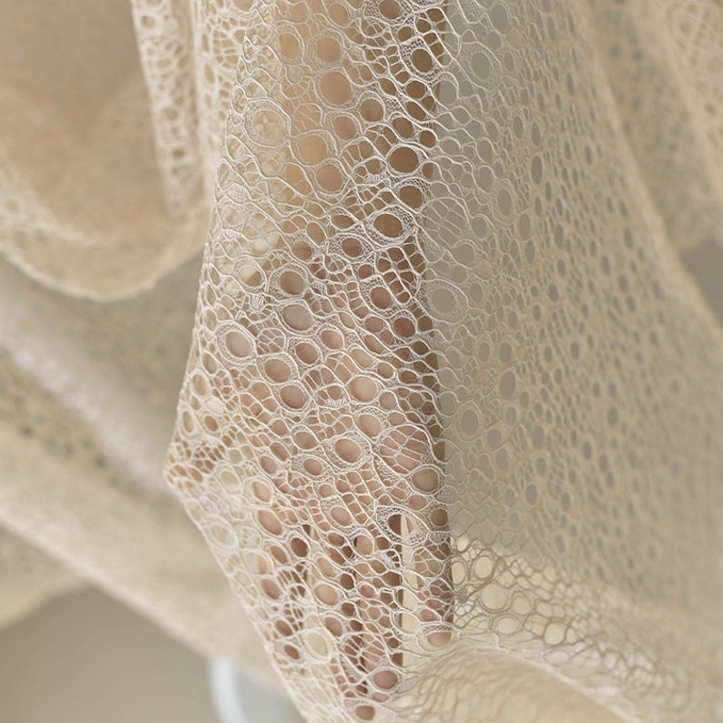 May include: Close-up of a beige lace fabric with a delicate, circular pattern. The fabric is soft and flowing, with a subtle sheen.