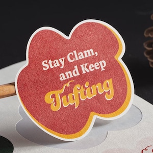 May include: A red, cloud-shaped sticker with a yellow border featuring the white and yellow text Stay Clam, and Keep Tufting. The sticker has a textured surface and is attached to a wooden stick, positioned against a light-coloured paper backing.
