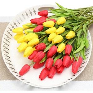 Op de afbeelding: Een boeket kunstmatige tulpen in rood en geel. De tulpen zijn gerangschikt in een witte schaal met een decoratieve rand.