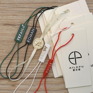 Puede incluir: Una colección de etiquetas colgantes rectangulares de color blanco roto con cordones. Las etiquetas incluyen cierres de plástico en verde, negro, blanco y rojo, impresos con nombres de marca como PKPAOLO, ROMAN y AILEPU, y un emblema dorado circular.
