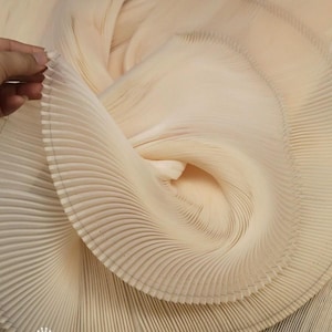 May include: A light beige fabric featuring dense, uniform accordion pleats that create a swirling, circular pattern. The material is lightweight and semi-translucent, with visible stitching along the edges of the pleats forming a structured, layered texture.